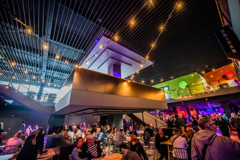 Een drukke moderne binnenruimte met meerdere niveaus, levendig verlicht en gevuld met mensen die aan tafels zitten en socialiseren.