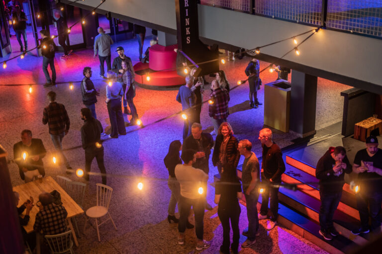 Een bovenaanzicht van een levendig evenement met mensen die in groepen staan, omringd door sfeervolle verlichting in verschillende kleuren.