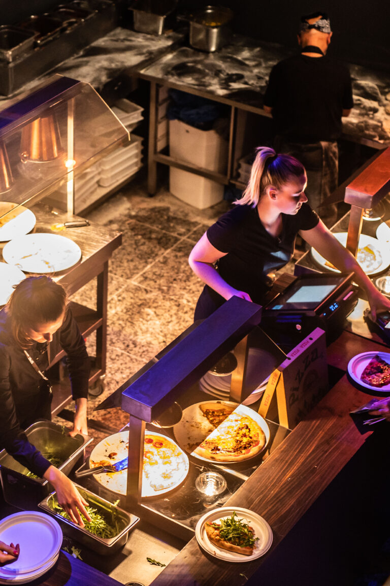Zicht op een buffet met twee medewerkers die gerechten voorbereiden en serveren in een sfeervolle omgeving.