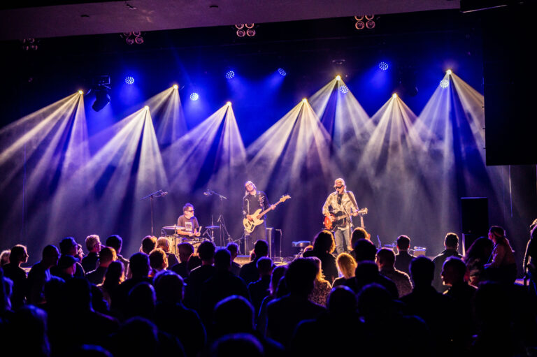 Een live band speelt op een podium met kleurrijke verlichting, terwijl een menigte toekijkt.