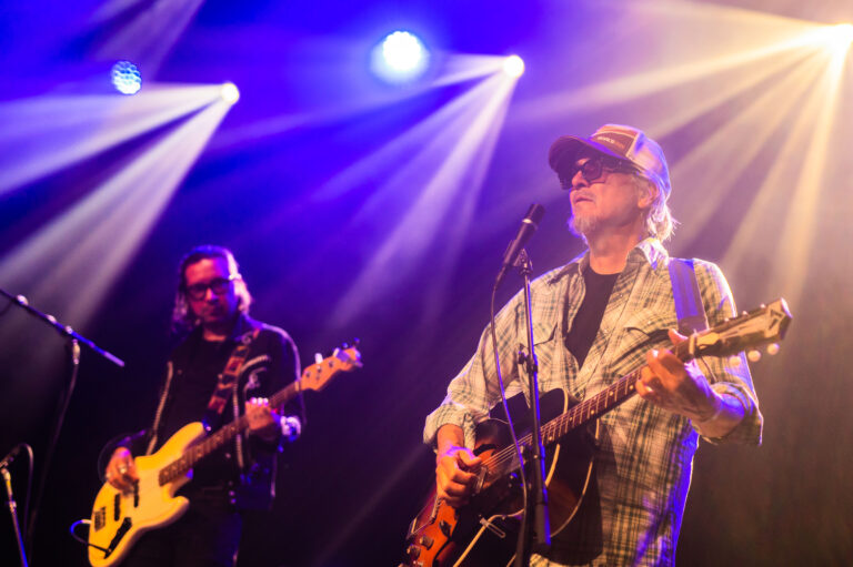 Een muzikant met een gitaar optredend voor een publiek, terwijl een bassist op de achtergrond speelt, verlicht door sfeervolle lichten.