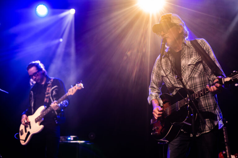 Twee muzikanten treden op het podium op met een gitaar en een basgitaar, verlicht door blauwe en gouden lichten.