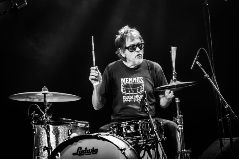 Een drummer met een zonnebril speelt enthousiast op zijn drumset tijdens een liveoptreden.