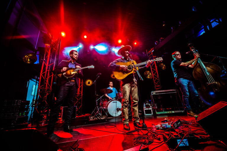 Een band speelt live muziek op een podium, omringd door felrode lichten en instrumenten.