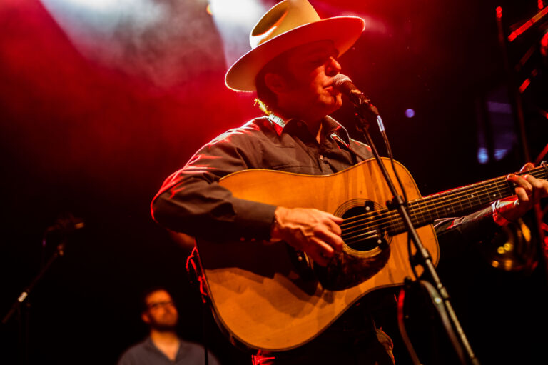 Een muzikant speelt gitaar en zingt op een podium met rode verlichting.