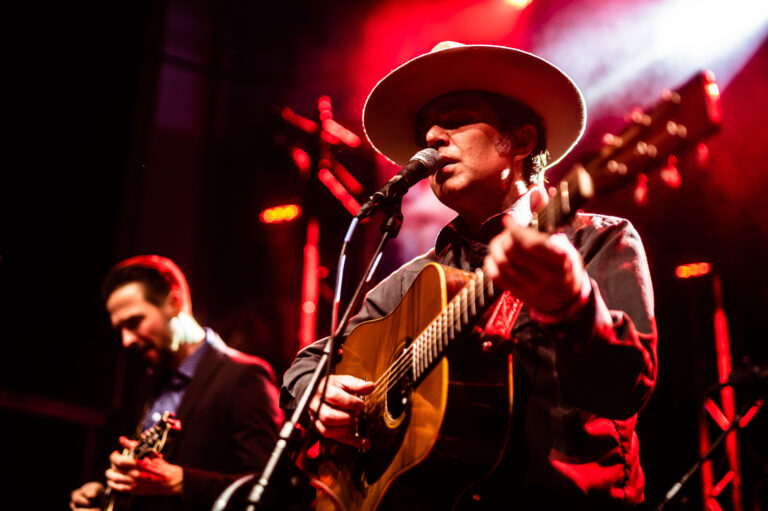 Een man met een hoed speelt gitaar en zingt op een podium met sfeervolle verlichting, terwijl een muzikant achter hem een mandoline bespeelt.