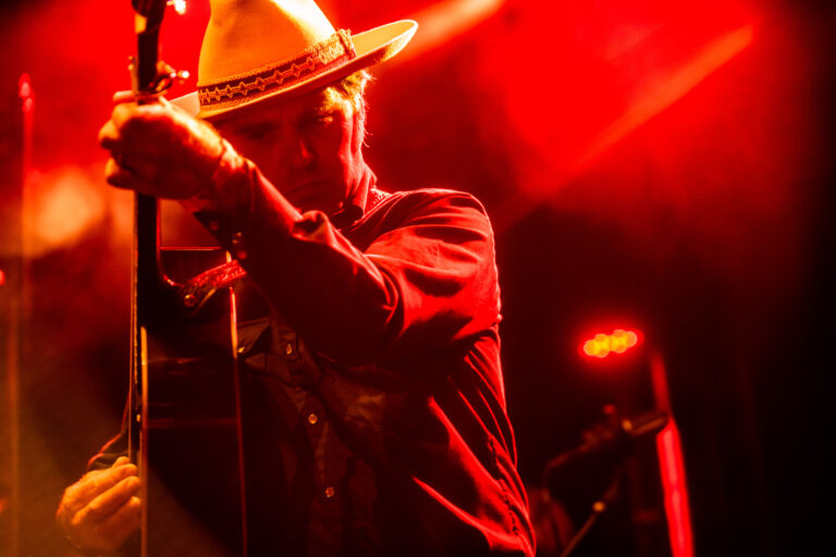 Een muzikant met een hoed speelt gitaar onder felrode lichtstralen op een podium.
