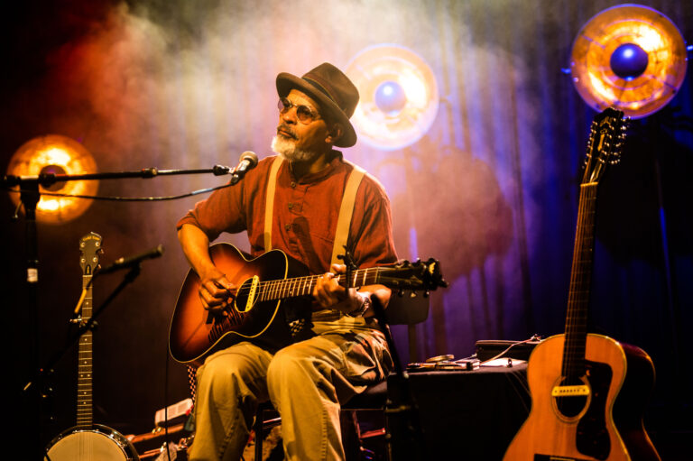 Een muzikant met een hoed speelt gitaar op een podium, omringd door sfeervolle verlichting.