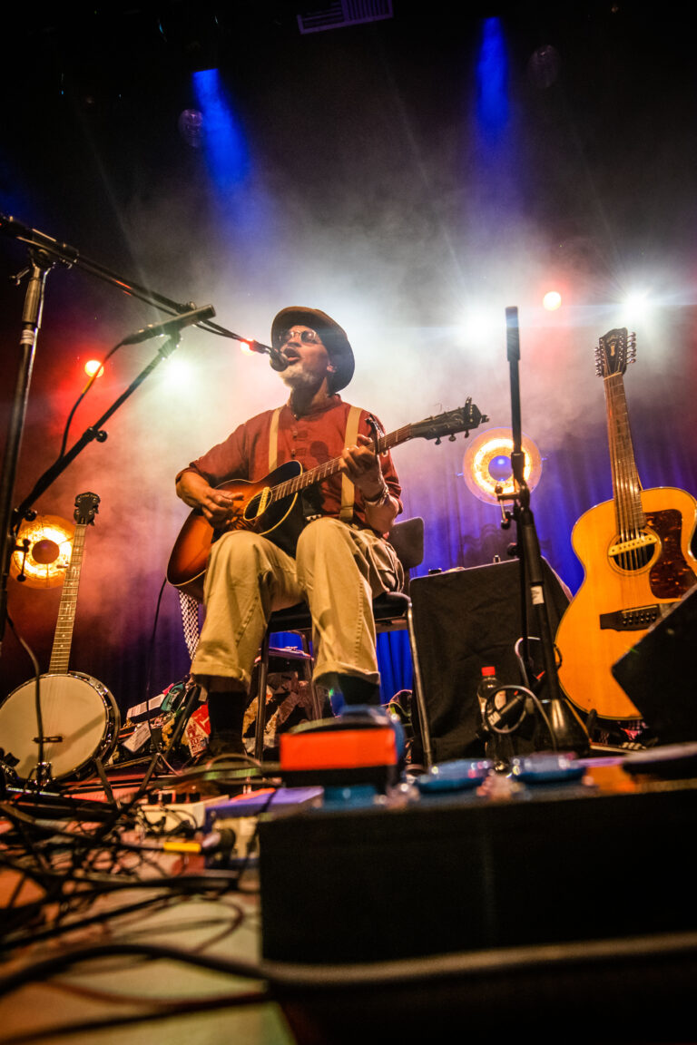Een muzikant zit op een stoel met een gitaar, omringd door instrumenten en sfeervolle verlichting op het podium.