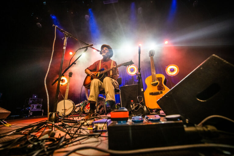 Een muzikant zit op het podium met een gitaar, omringd door instrumenten en kleurrijke lichten.