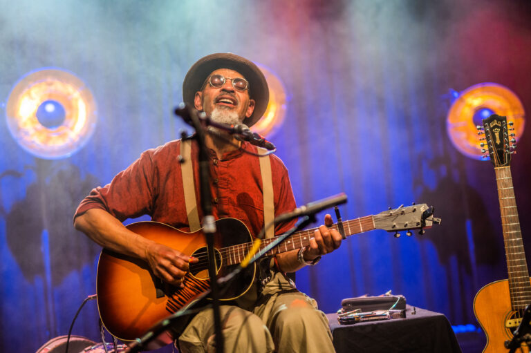 Een man speelt gitaar en zingt op het podium, omringd door sfeerverlichting.
