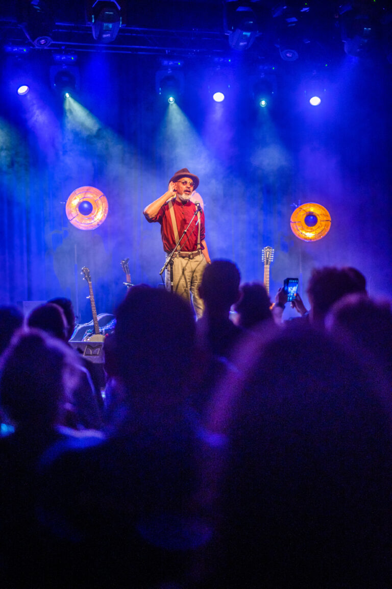 Een performer staat op een podium, omringd door een publiek en verlichte achtergrondlichten.