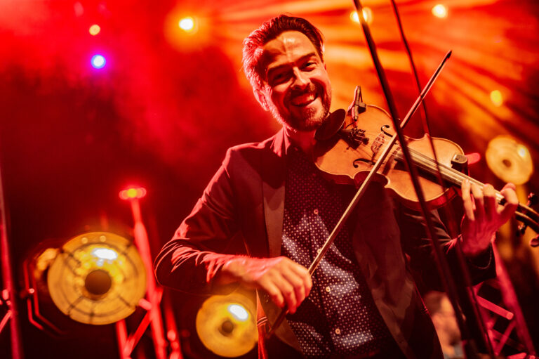 Een muzikant speelt vrolijk viool op een podium, omgeven door felgekleurde lichten.