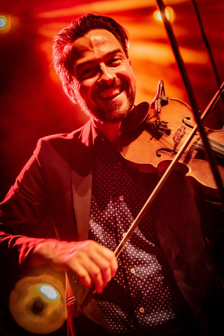 Een glimlachende man in een zwart jasje houdt een viool vast op een podium met een warme, vurige achtergrondverlichting.