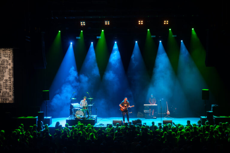 Een muzikant optredend op een podium, omringd door kleurrijke verlichting en een enthousiaste menigte.