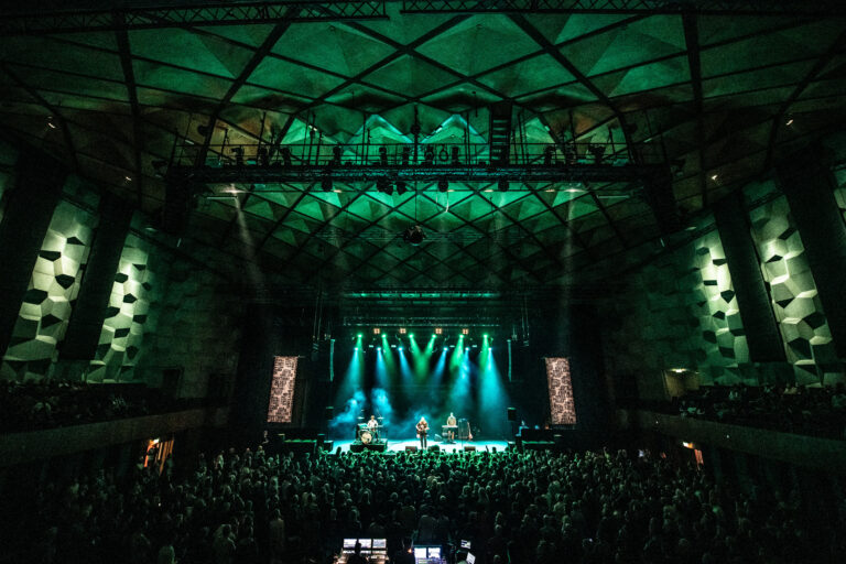 Een drukbezocht concert in een grote hal met groene verlichting en een hoge, unieke plafondstructuur.