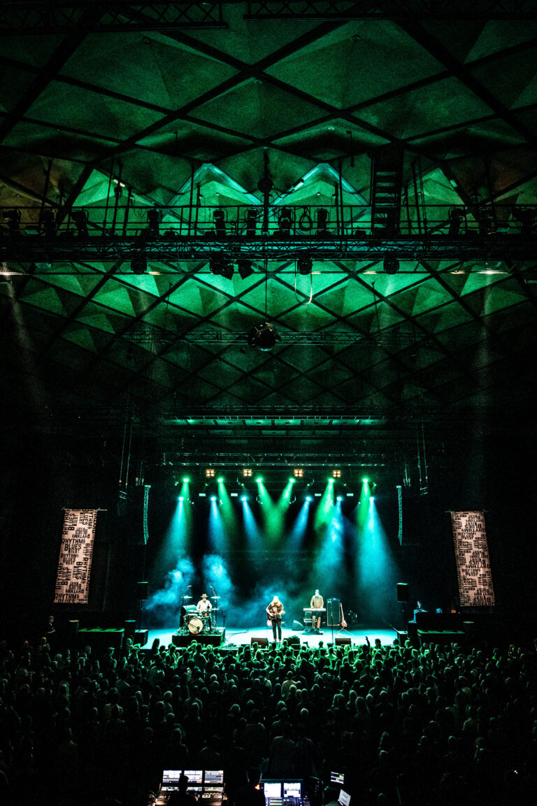 Een liveconcert met een band op het podium, omgeven door een groot publiek en verlicht door groene lichten in een donkere zaal.