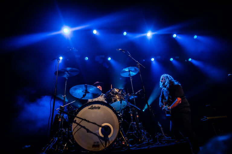 Een muzikant speelt gitaar naast een drumstel, verlicht door blauwe spotlights op een podium.