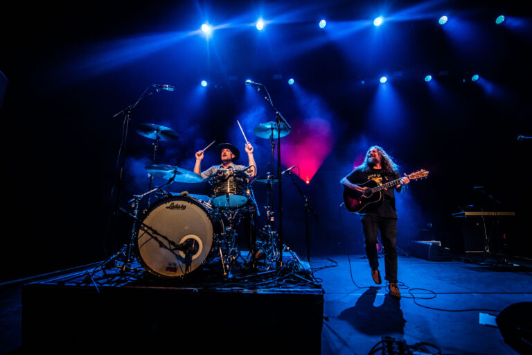 Een drummer en een gitarist spelen samen op het podium, verlicht door blauwe lichten.