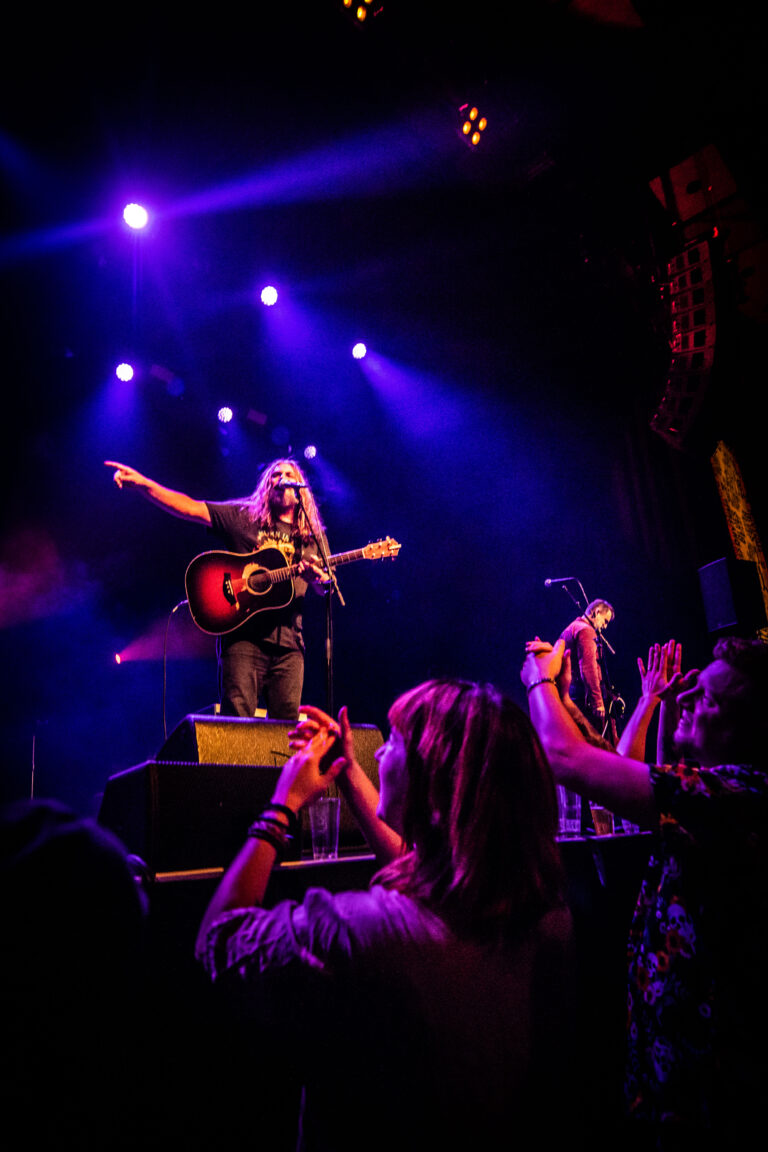 Een muzikant met een gitaar staat op het podium en zingt, terwijl het publiek enthousiast reageert op de performance.