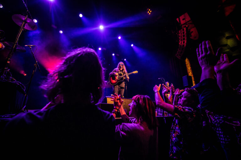 Een muzikant op een podium speelt gitaar terwijl het publiek enthousiast zijn handen opsteekt, omgeven door kleurrijke verlichting.