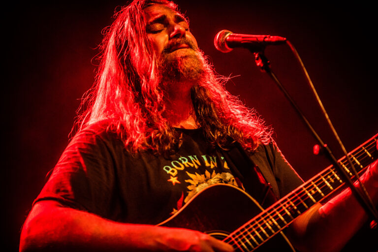 Een muzikant met lang haar zingt emotioneel in een lichteffect met rode tonen, terwijl hij een akoestische gitaar vasthoudt.