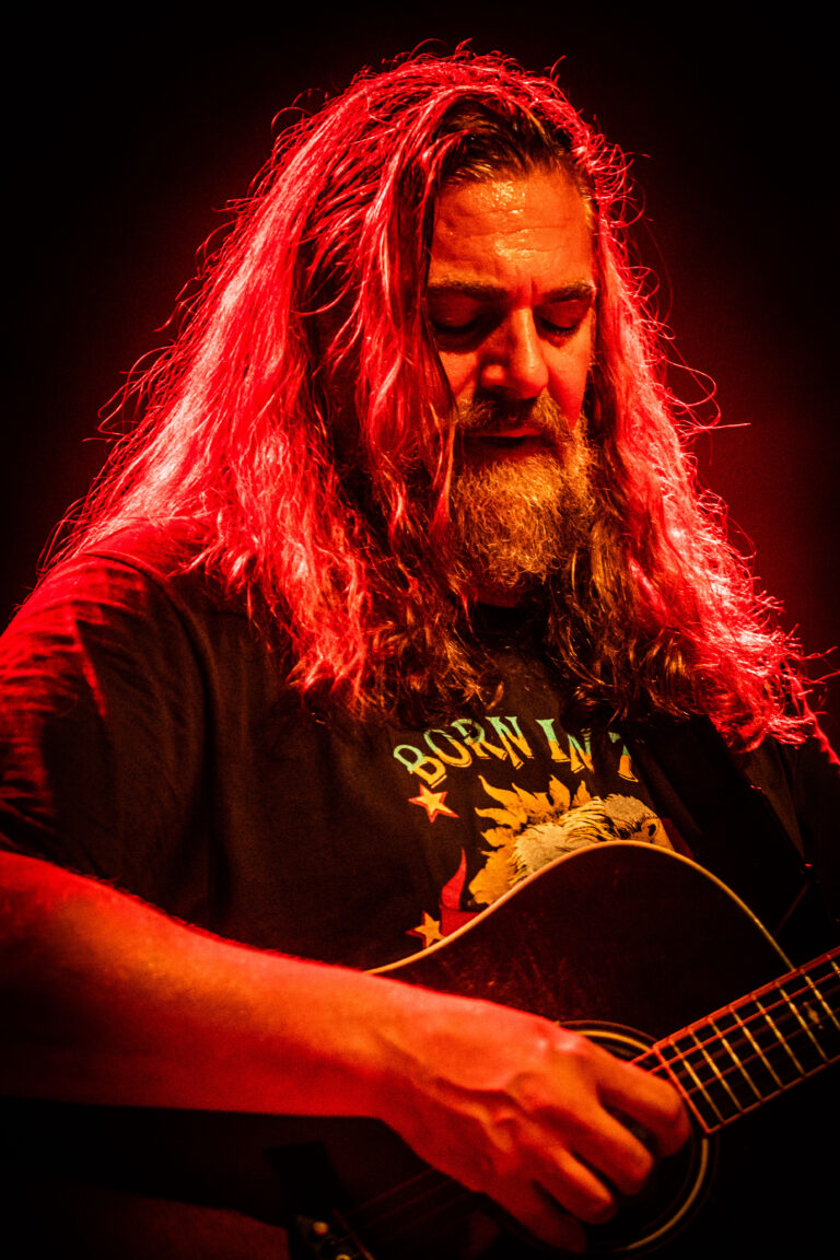 Een man met een volle baard en lang haar speelt gitaar onder een rode belichting.