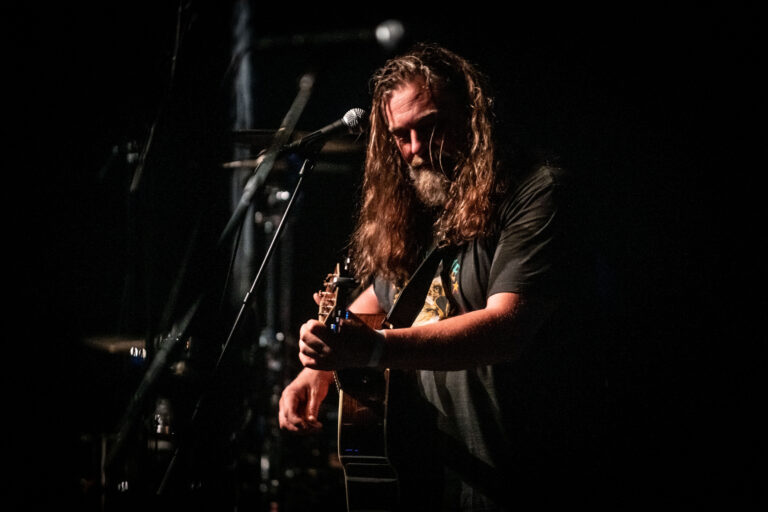 Een muzikant met lang haar speelt gitaar op een donkere podium, omringd door microfoons en andere instrumenten.