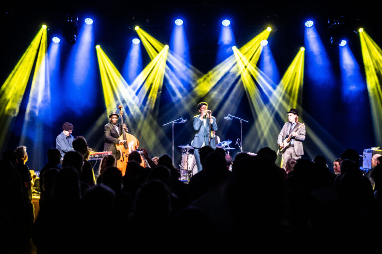 Een band speelt op een podium met dynamische verlichting en een menigte die geniet van de muziek.