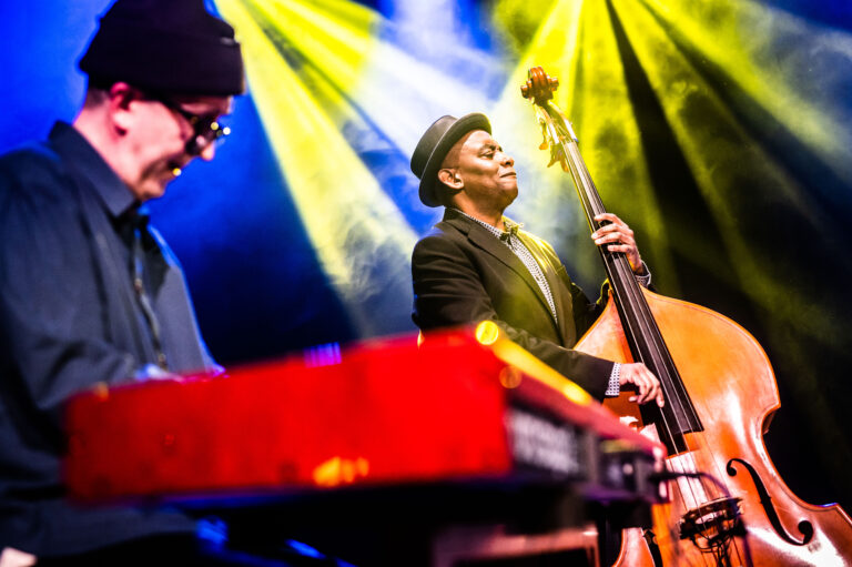 Twee muzikanten spelen samen op een podium, waarbij de bassist centraal staat met een contrabas en kleurrijke lichten achter hen stralen.