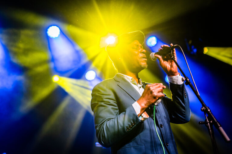 Een zanger staat op het podium met een microfoon, omringd door kleurrijke lichtstralen.