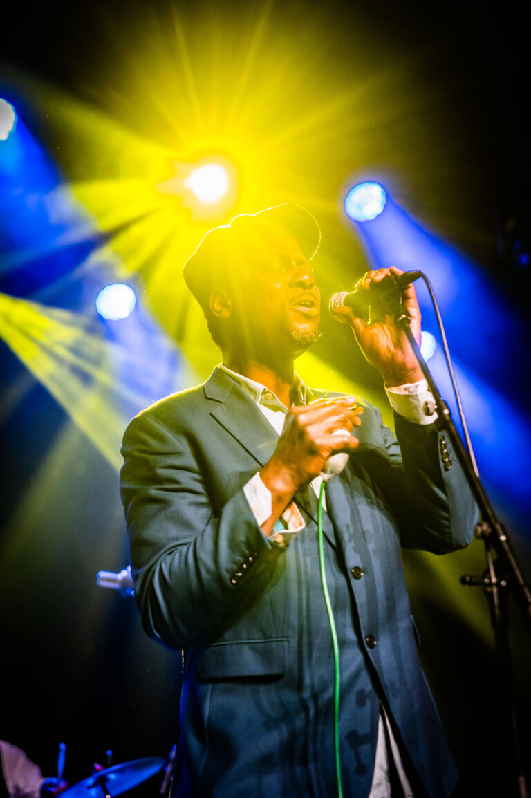 Een zanger in een blauwe blazer staat op het podium en zingt, omringd door kleurige spotlights.