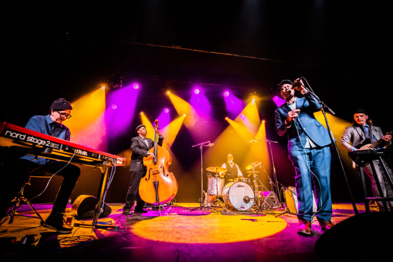 Een muzikale band staat op het podium met verschillende instrumenten, omgeven door kleurrijke verlichting.