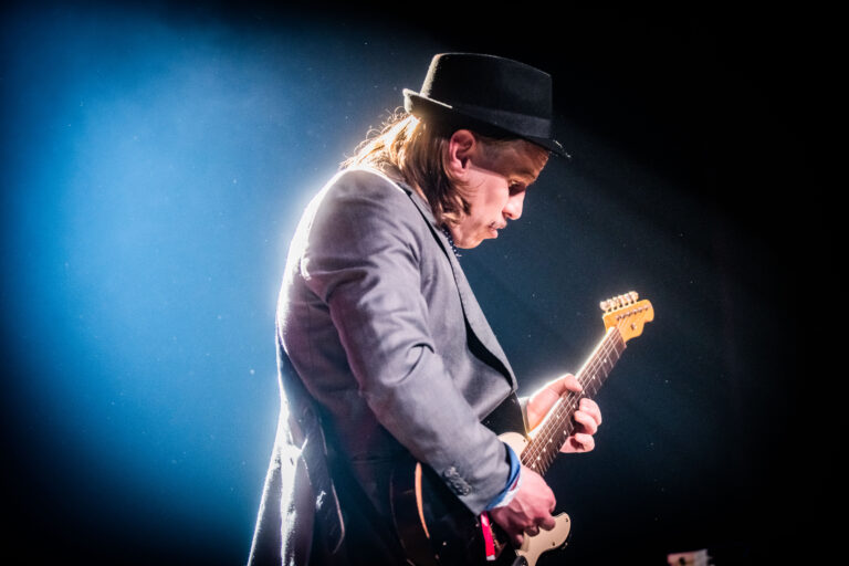 Een muzikant speelt gitaar op het podium met een hoed en een grijs jasje, omringd door een felblauwe achtergrond.