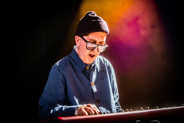 Een muzikant speelt op een keyboard, gekleed in een blauwe blouse en een muts, onder een kleurrijke achtergrond.