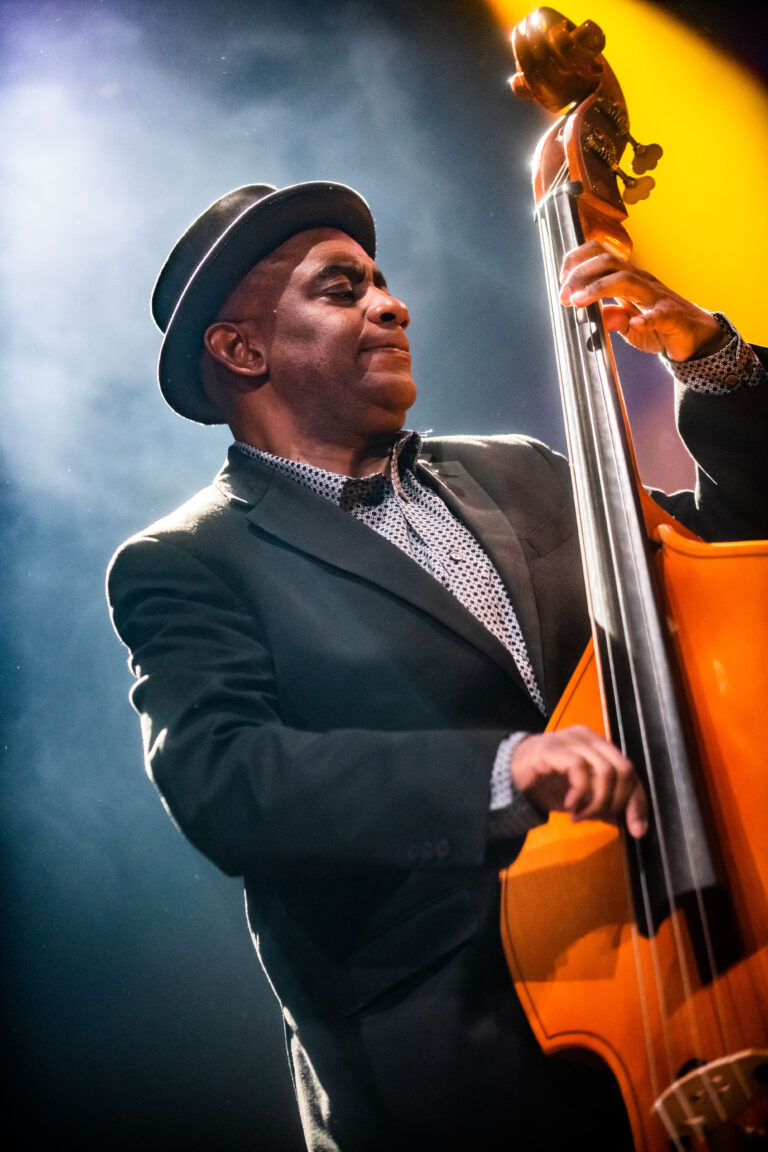 Een muzikant speelt een contrabas op een podium, omringd door kleurrijke lichtstralen.