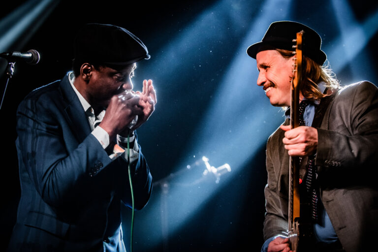 Twee muzikanten treden op, waarbij de ene een mondharmonica bespeelt en de andere een elektrische gitaar vasthoudt, met theatrale verlichting op de achtergrond.
