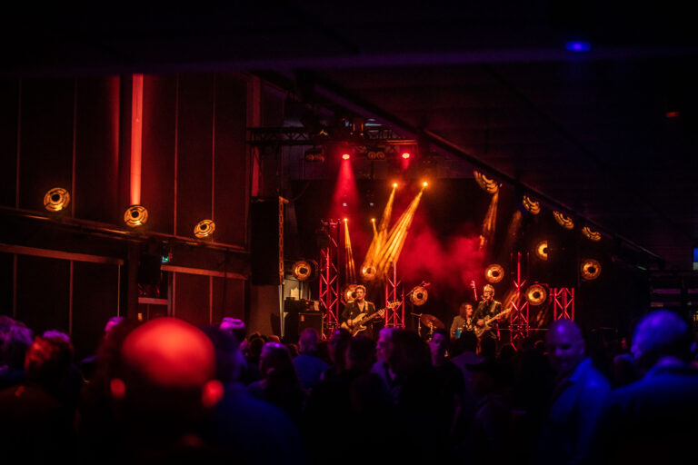 Een liveband speelt op een podium met kleurrijke verlichting, omringd door een enthousiaste menigte.
