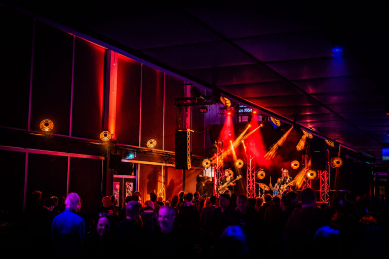 Optreden van een band met kleurrijke verlichting en een enthousiaste menigte in een binnenlocatie.