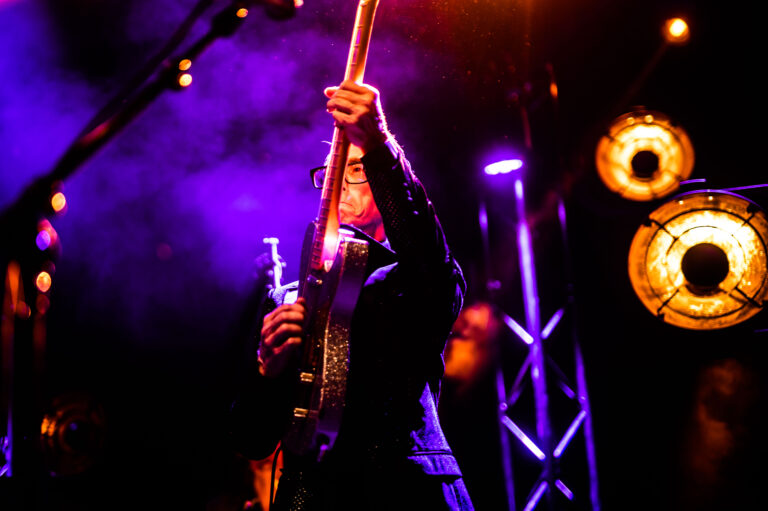 Een muzikant speelt gitaar op een podium, omringd door kleurrijke verlichting en rook.