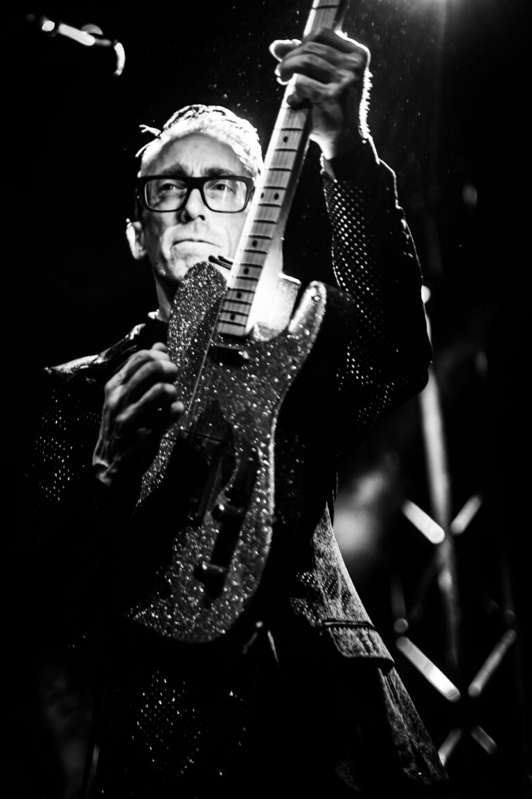 Een muzikant in een glinsterend zwart kostuum houdt een gitaar vast terwijl hij op het podium staat.