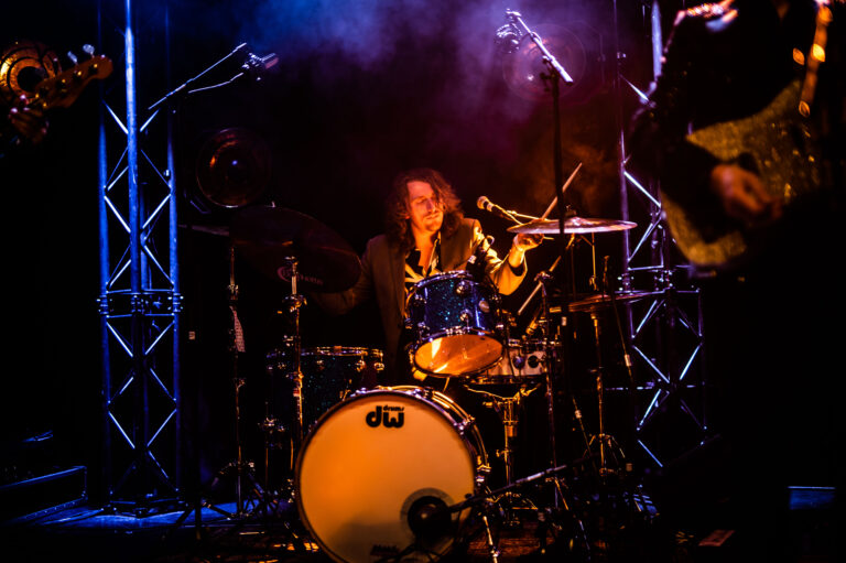Een drummer speelt op een podium, omringd door kleurrijke verlichting en rook.