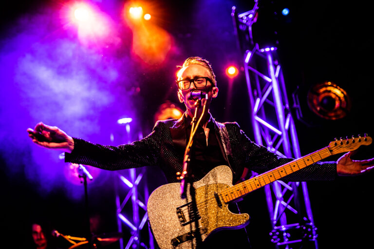 Een muzikant met een gitaar staat op het podium, omringd door kleurrijke lichteffecten.