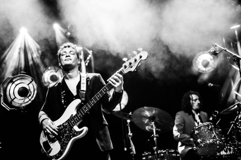 Een bassist speelt op het podium, terwijl een drummer op de achtergrond meedoet, omgeven door helder licht en rook.