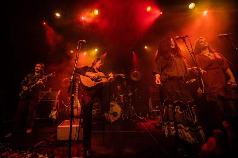 Een band speelt live op het podium, omringd door kleurrijke verlichting en rook.