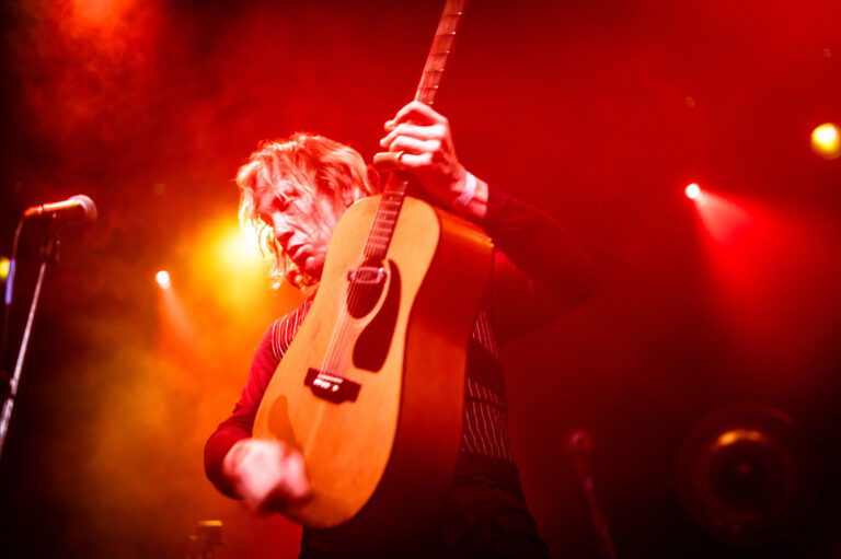 Een muzikant speelt een akoestische gitaar op een podium met kleurrijk licht.