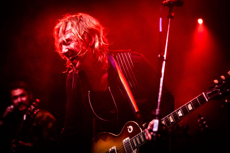 Een muzikant speelt energiek gitaar op het podium, omgeven door een roodgekleurde lichtshow.