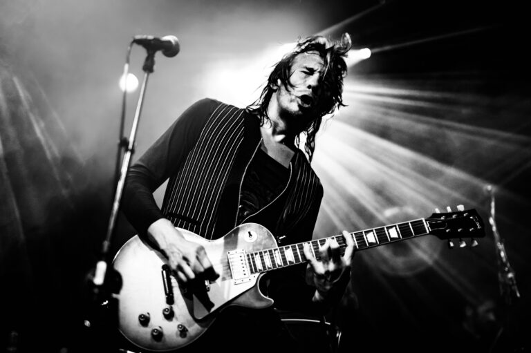 Een muzikant speelt gitaar op een podium met lichtstralen die eromheen schijnen.