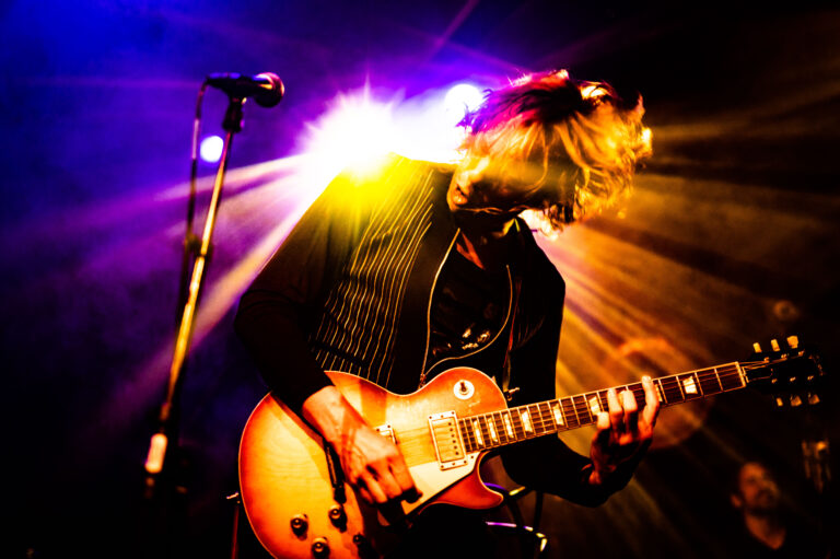 Een muzikant speelt gefocust op een elektrische gitaar met kleurrijke verlichting op de achtergrond.