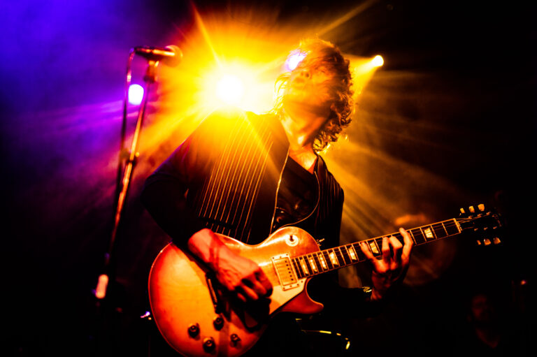 Een muzikant speelt gitaar op een podium met kleurrijke lichten en een sterke straling achter hen.